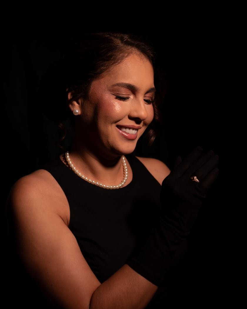 Suelândia sorrindo, de vestido preto e colar de pérolas, com um anel na mão